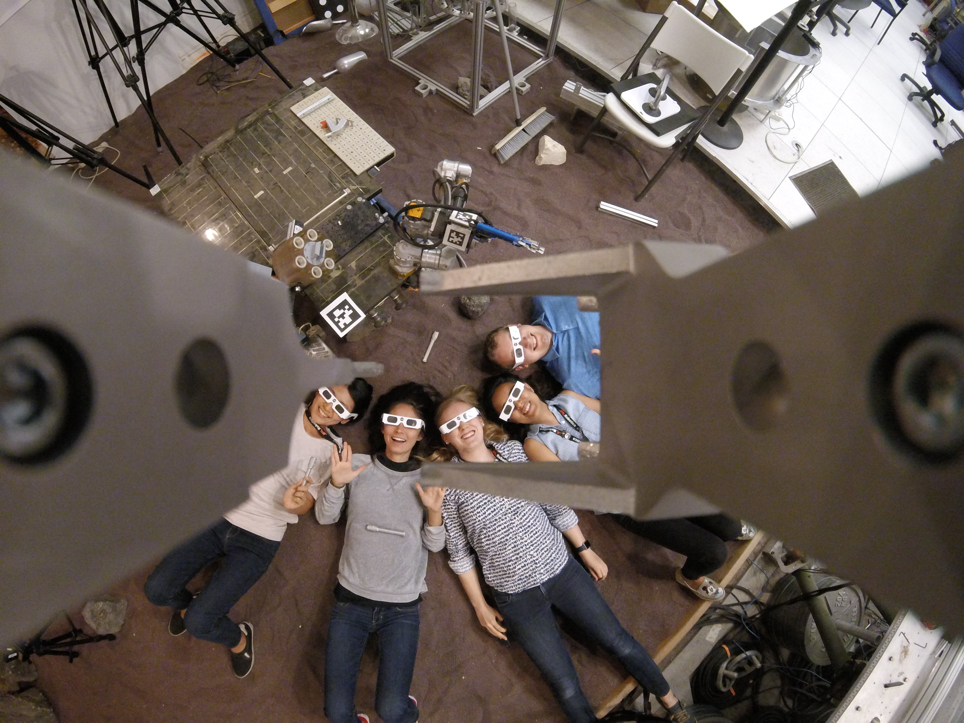 A group of male and female interns at NASA's Jet Propulsion Laboratory pose for photos in eclipse glasses. Looking up in a circle the group smiles and wears business casual attire laboratory instruments and devices are visible around the peripheral of the image. Credit: NASA/JPL-Caltech