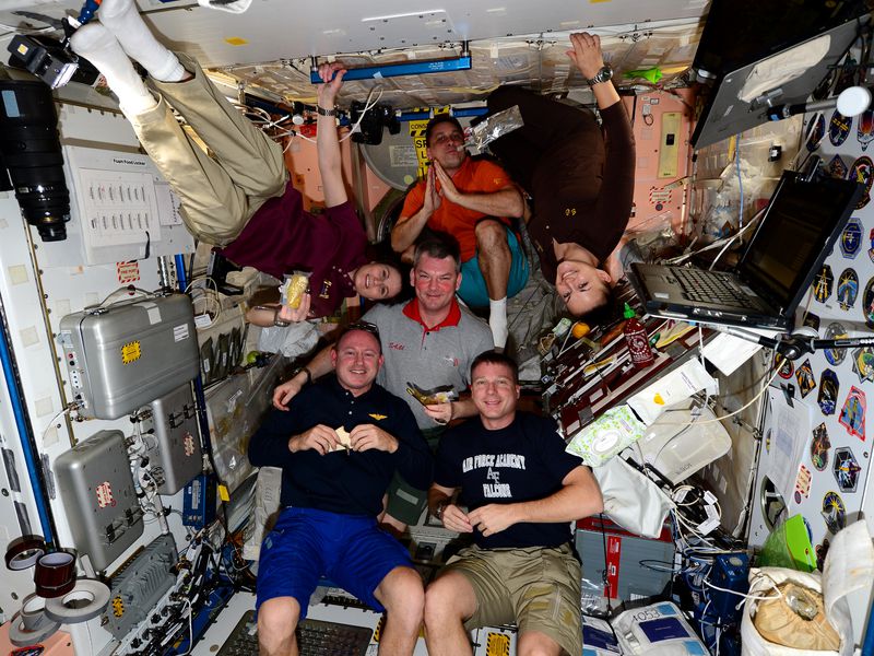 Three astronauts wearing black shirts, one astronaut wearing a burgundy shirt, one astronaut wearing a grey shirt with a red collar, and one astronaut wearing an orange shirt are all gathered together in a small space onboard NASA's International Space Station. Two astronauts (one in a burgundy shirt and one in a black shirt) are floating upside down. They are all eating.