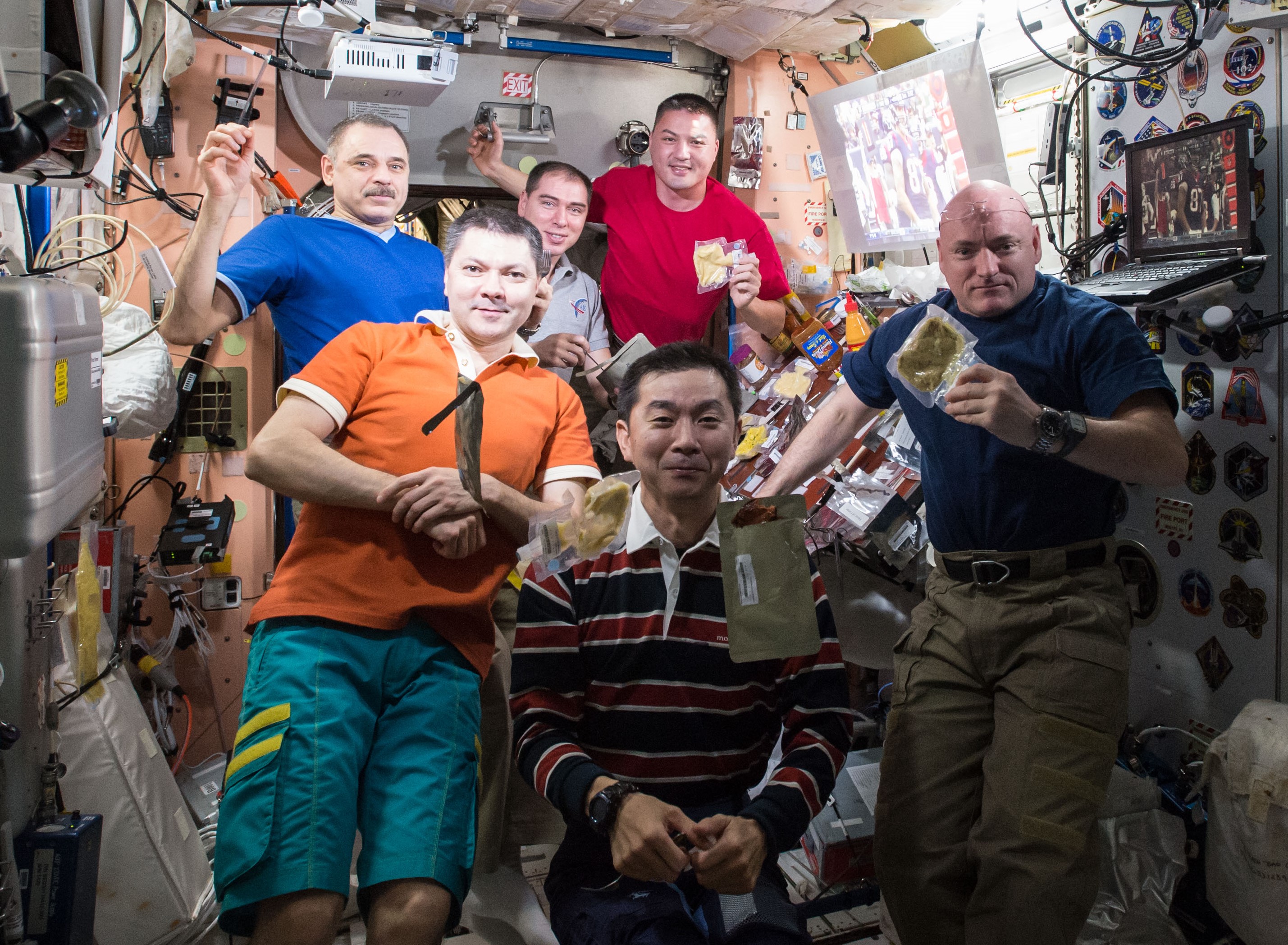 Six astronauts are gathered together onboard NASA's International Space Station. Vaccuum sealed bags of food are floating in the air. The left and right walls, and ceiling, is visible. Wires, a laptop. and stickers are attached to the walls. The stickers are of various designs meant to reflect the patches for each crewed mission.