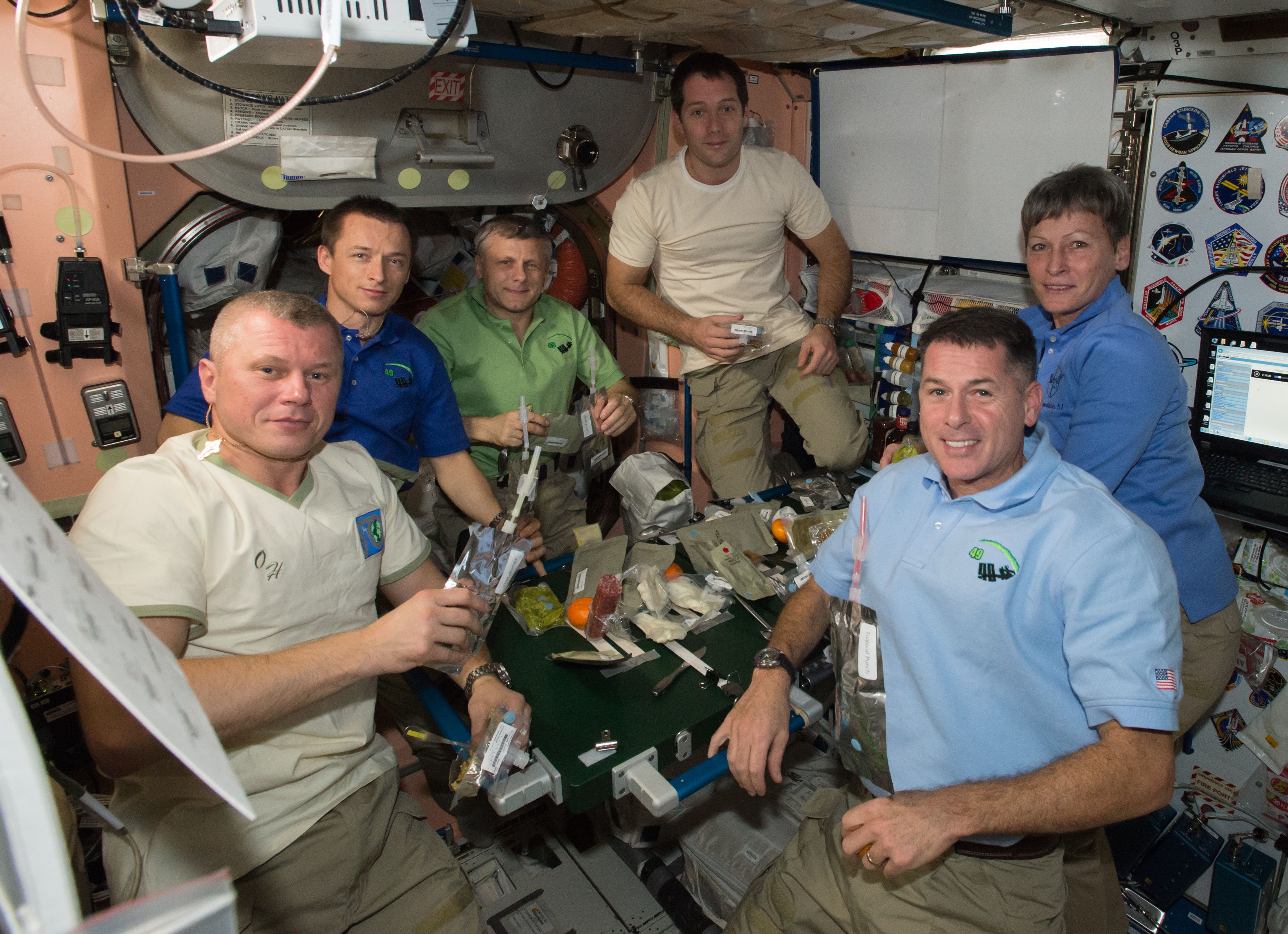 Six astronauts are gathered around a small table while onboard a spacecraft. They are holding various food items and smiling into the camera.