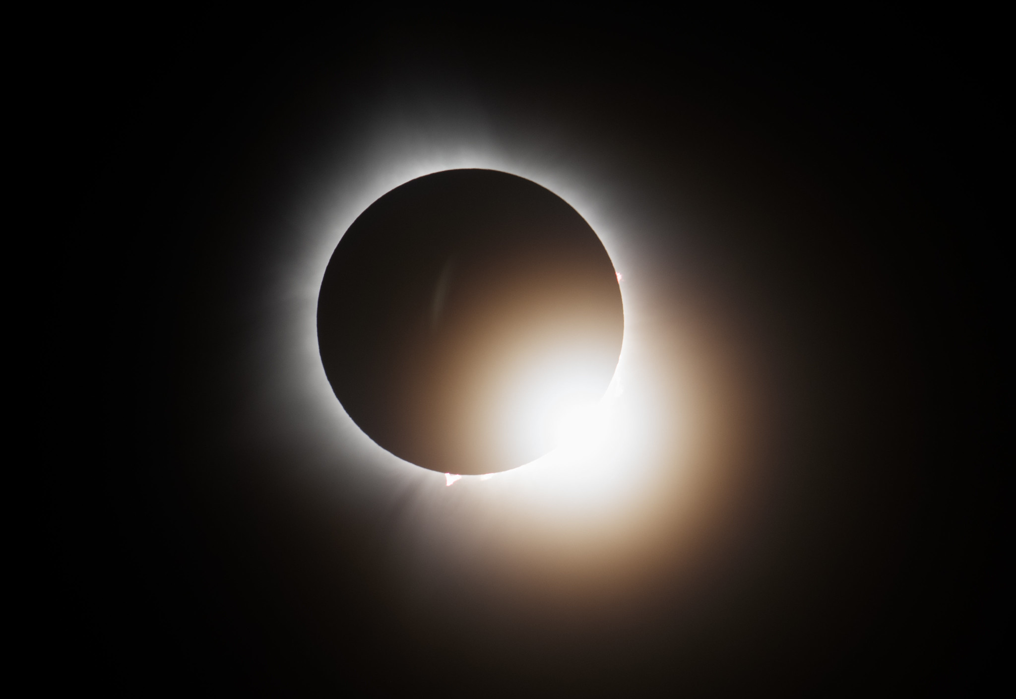 The total solar eclipse. Against a black background, there is a faint white glow surrounding a black circle. On the bottom right of this circle is a bright glowing circle overlapping.