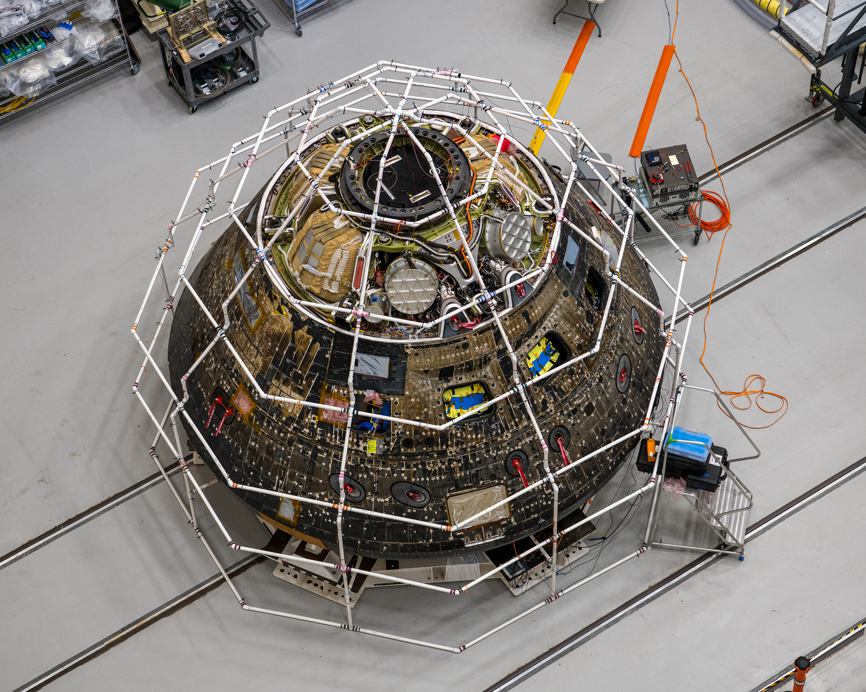 The Orion crew module pictured from overhead in the Space Environment Complex Assembly High Bay. Many of the vehicle covers were removed, and wires and hardware exposed. White PVC pipe covers the vehicle like a cage.