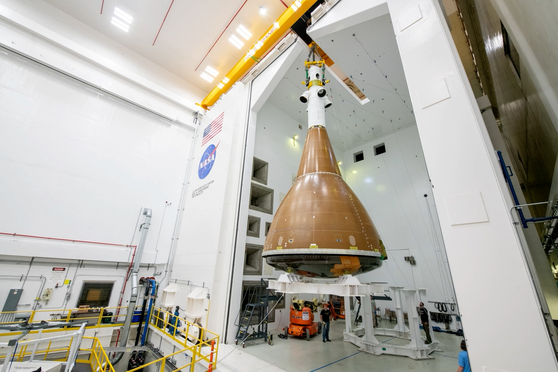 A yellow crane lifts the Orion crew module with launch abort system attachment into the Reverberant Acoustic Test Facility. The test hardware is a brownish orange blunted conical shape with a white cylinder shape at the top. The white walls are covered with large gray inset horns. A man wearing glasses, blue jeans, and a black t-shirt stands beneath the white platform that the test article is sitting on. The American flag and NASA logo appear on the wall to the left of the open facility door. [PHOTO – GRC-2024-C-13339.jpg]