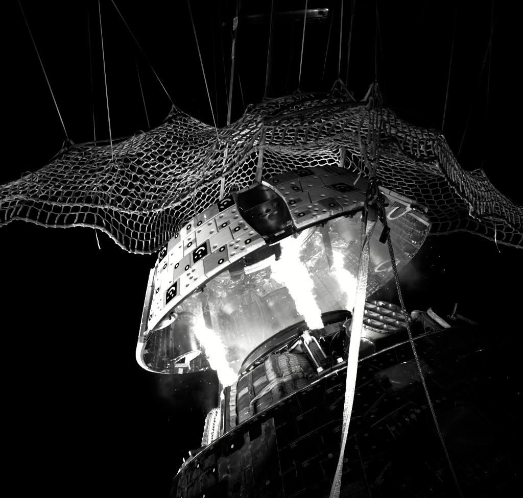 A black and white still image of the forward bay cover ejecting from the Orion crew module. Three bursts of flame jettison the cover upwards into a net.