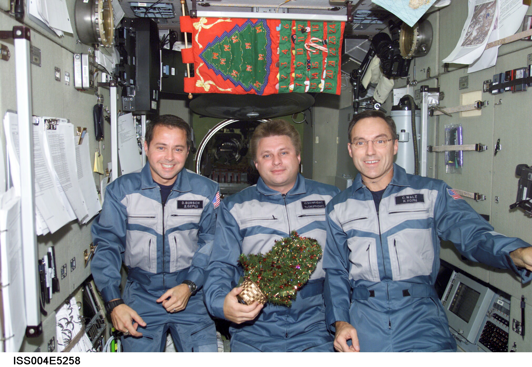 Three men in blue/gray suits smile as they pose for a Christmas pose onboard the Interrnational Space Station