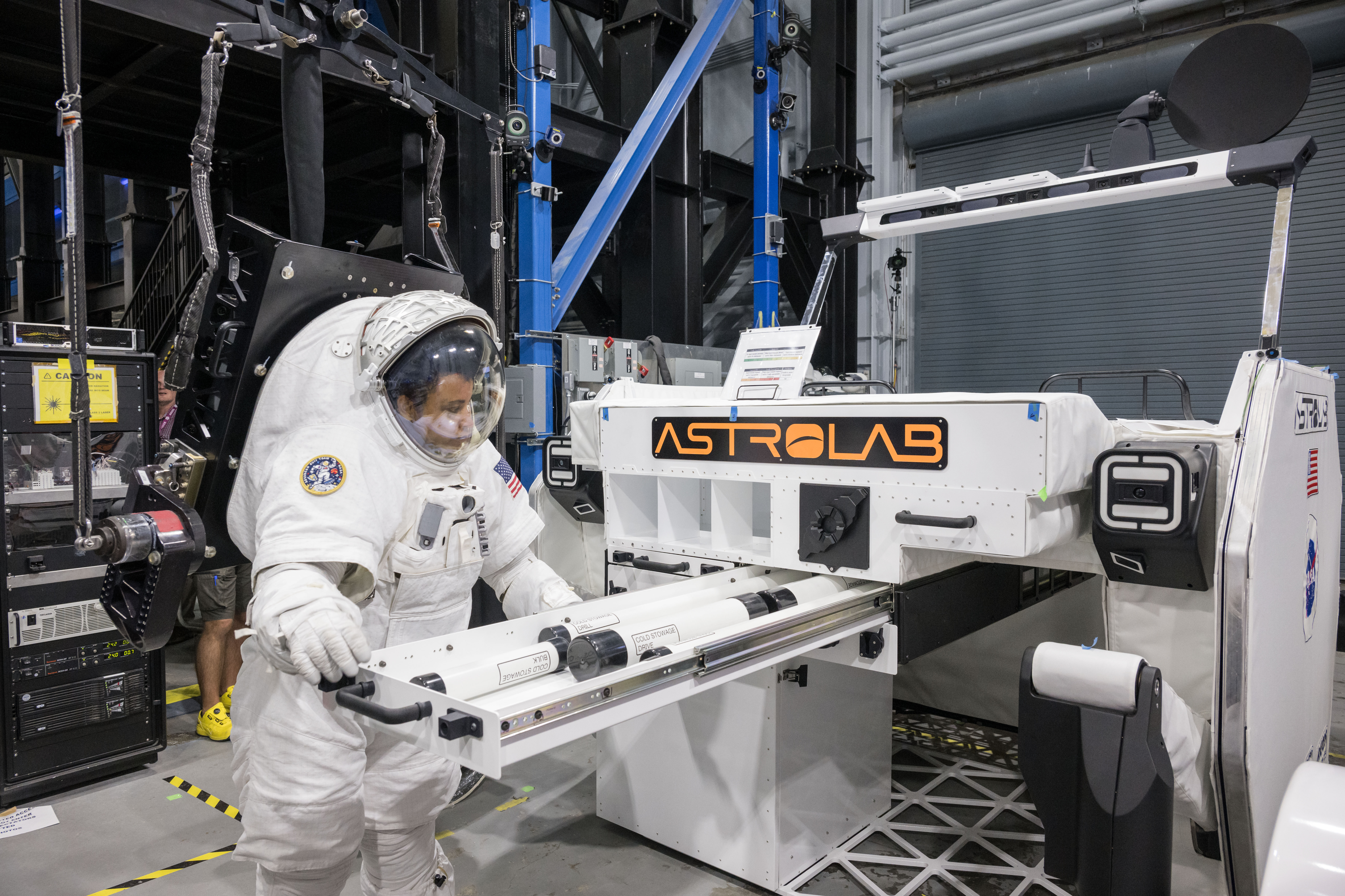 NASA astronaut Jessica Watkins is wearing a white spacesuit while she puts science payloads in a stowage drawer outside of Astrolab's white lunar terrain vehicle. There is an orange Astrolab logo on the rover.