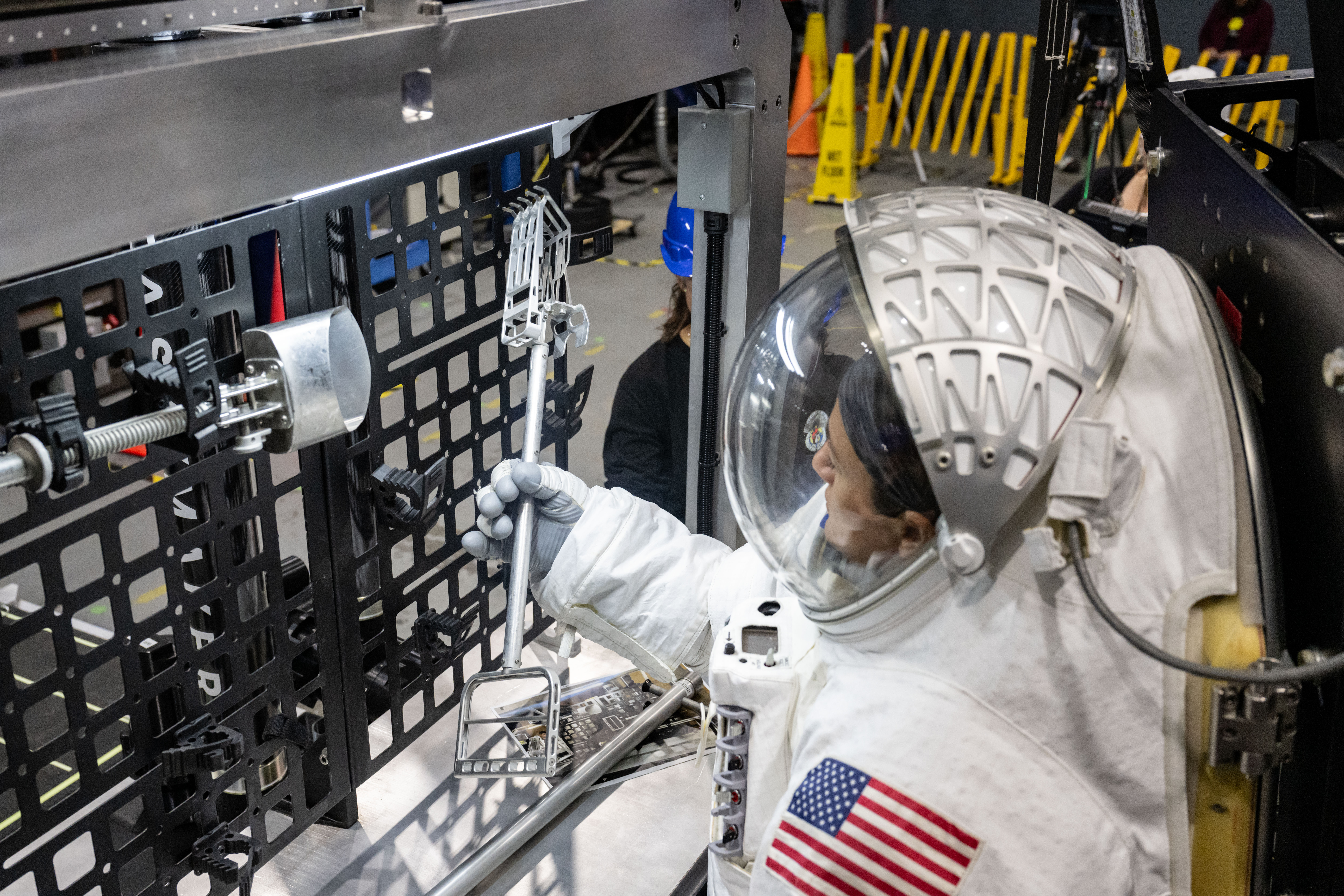 NASA astronaut Jessica Meir is shown in a white spacesuit with the American flag on her left shoulder as she grabs a lunar geology tool from a tool rack on Lunar Outpost’s Eagle lunar terrain vehicle.