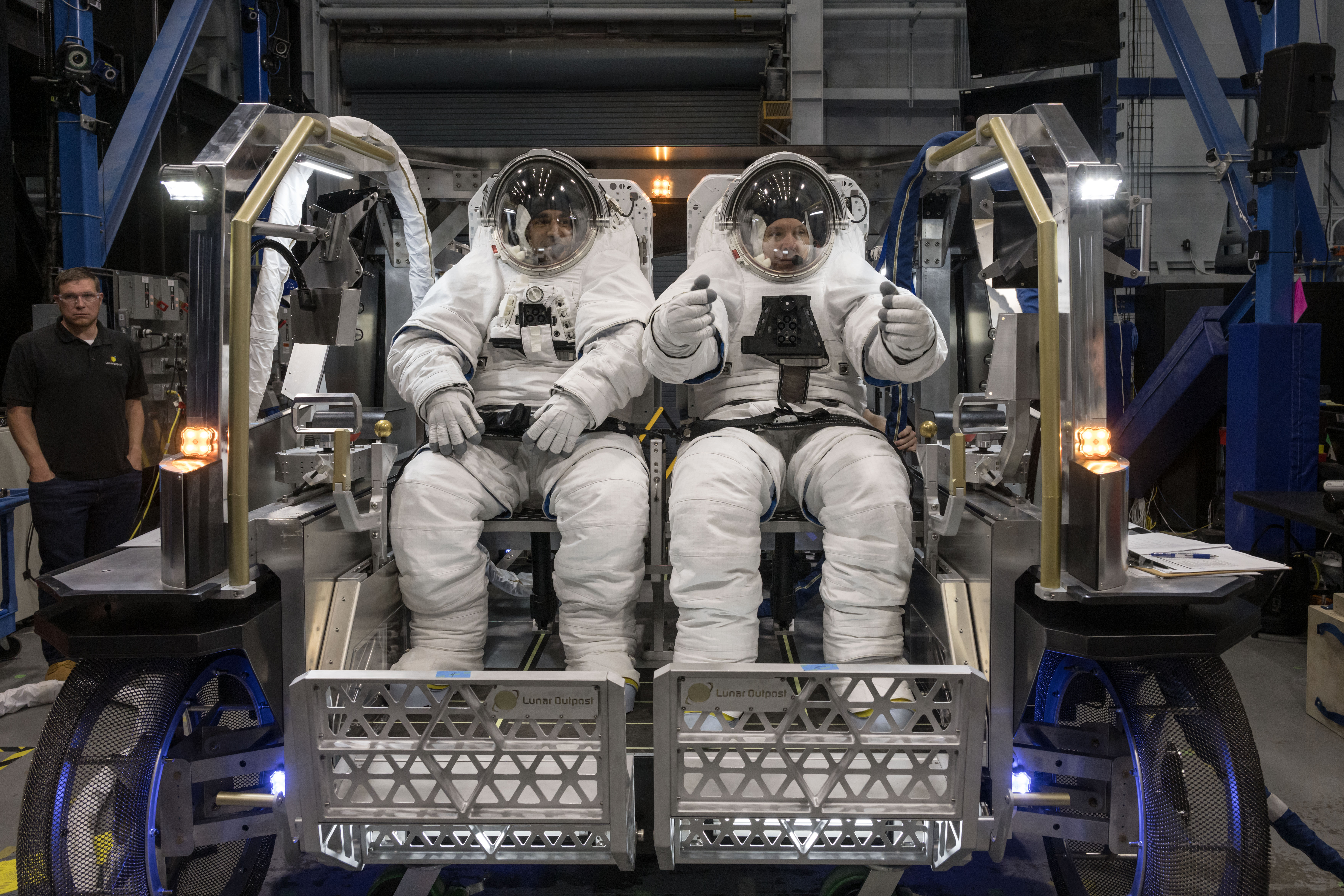 NASA astronauts Raja Chari and Randy Bresnik sit side by side inside Lunar Outpost’s Eagle lunar terrain vehicle while wearing white spacesuits.