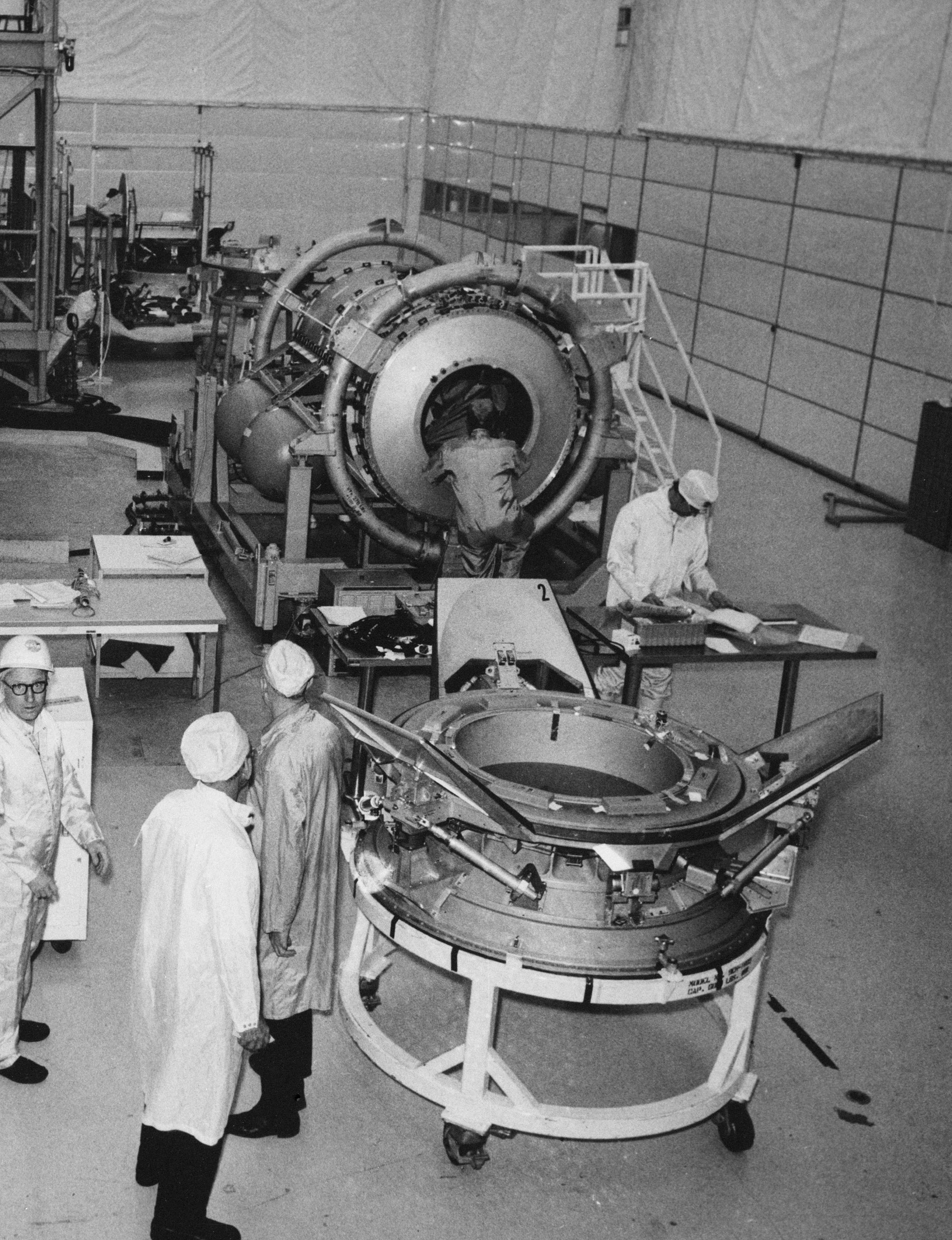 Black and white image of two spacecraft components with white coated technicians in a large room