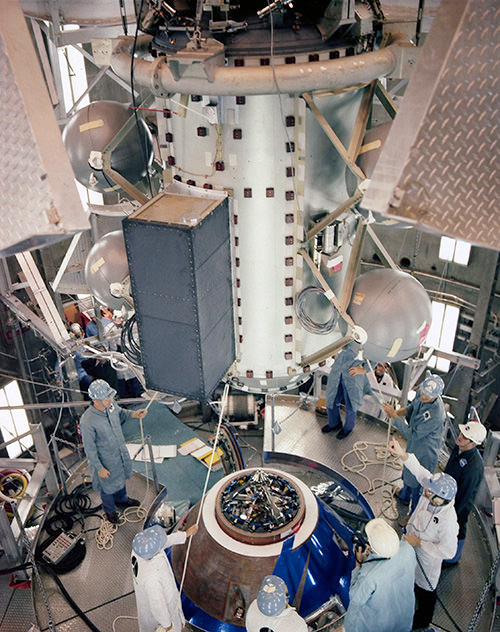 Workers lower a cylindrical spacecraft onto a cone shaped docking target in an altitude chamber