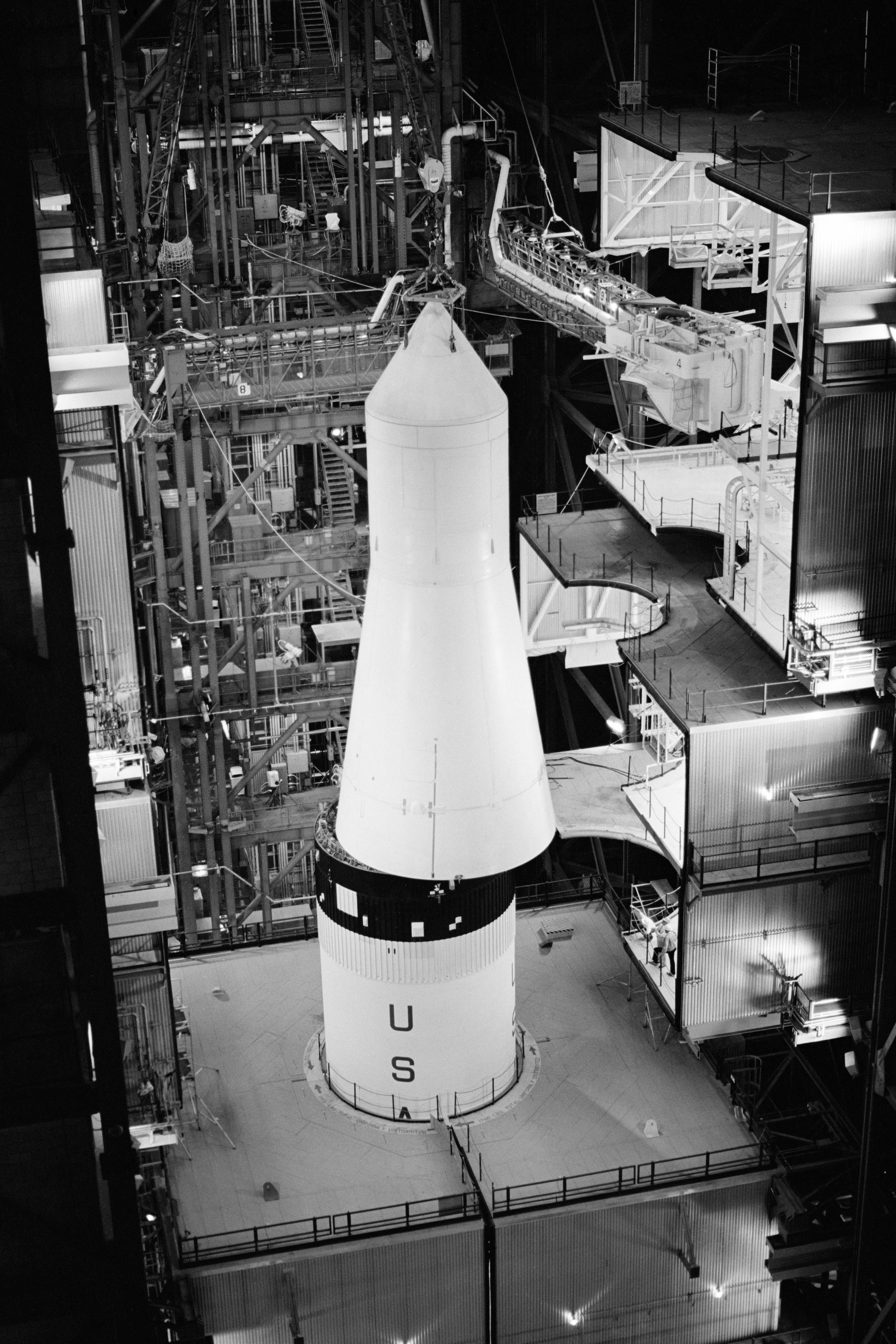 A white cone-shaped spacecraft is lifted and about to be placed atop a rocket inside a large assembly building