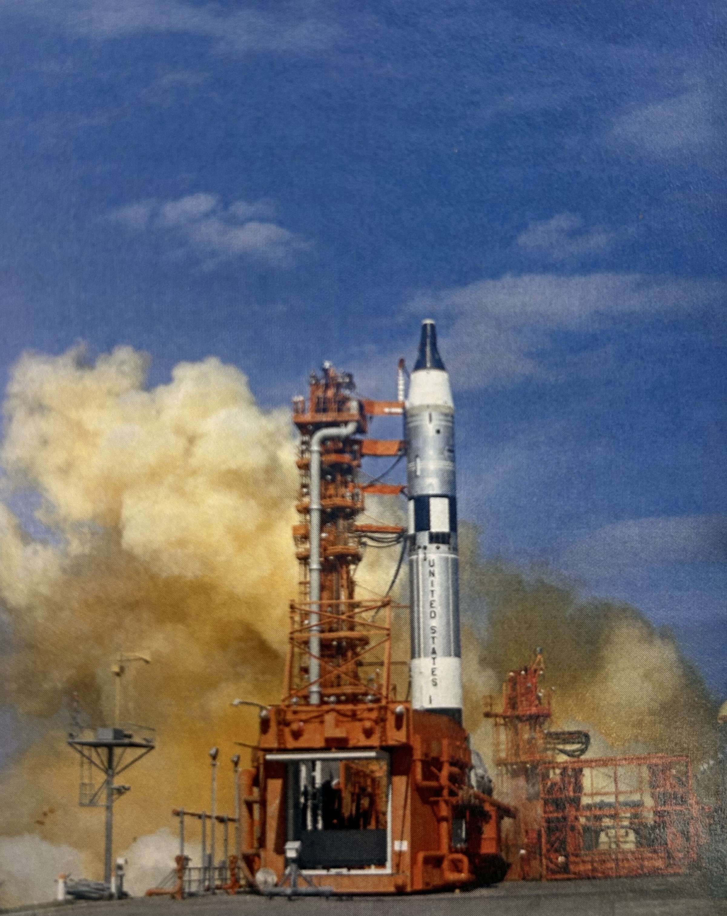 Image of a white, gray, and black rocket next to a read launch pad gantry, with an orange cloud of exhaust streaming to both sides of the roclet.