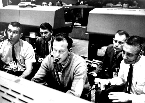 A group of five men in white shirts and ties hunched over a console in a control center.