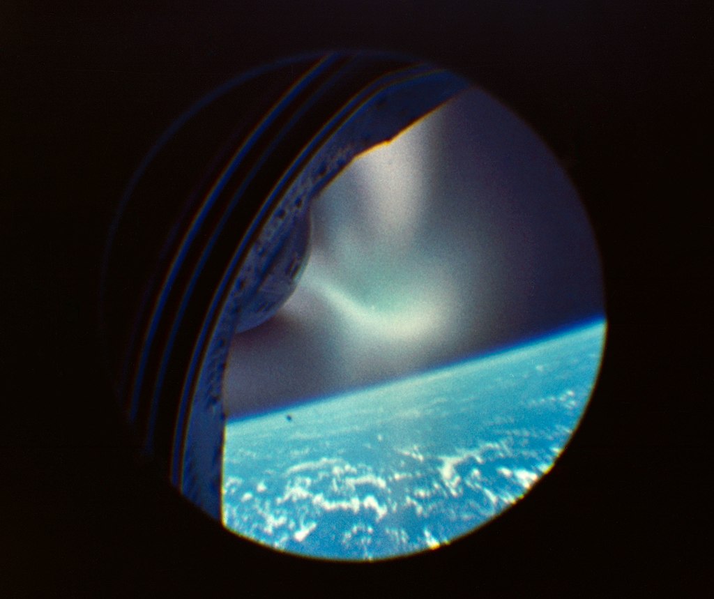 Image taken through the window of a spacecraft during reentry, showing wisps of flame against the blue limb of the Earth and the blackness of space.