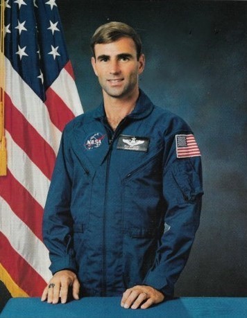 Portrait of a man wearing a blue jumpsuit with a partially visible US flag in the background.