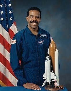 Portrait of a man wearing a blue jumpsuit with a partially visible US flag in the background.