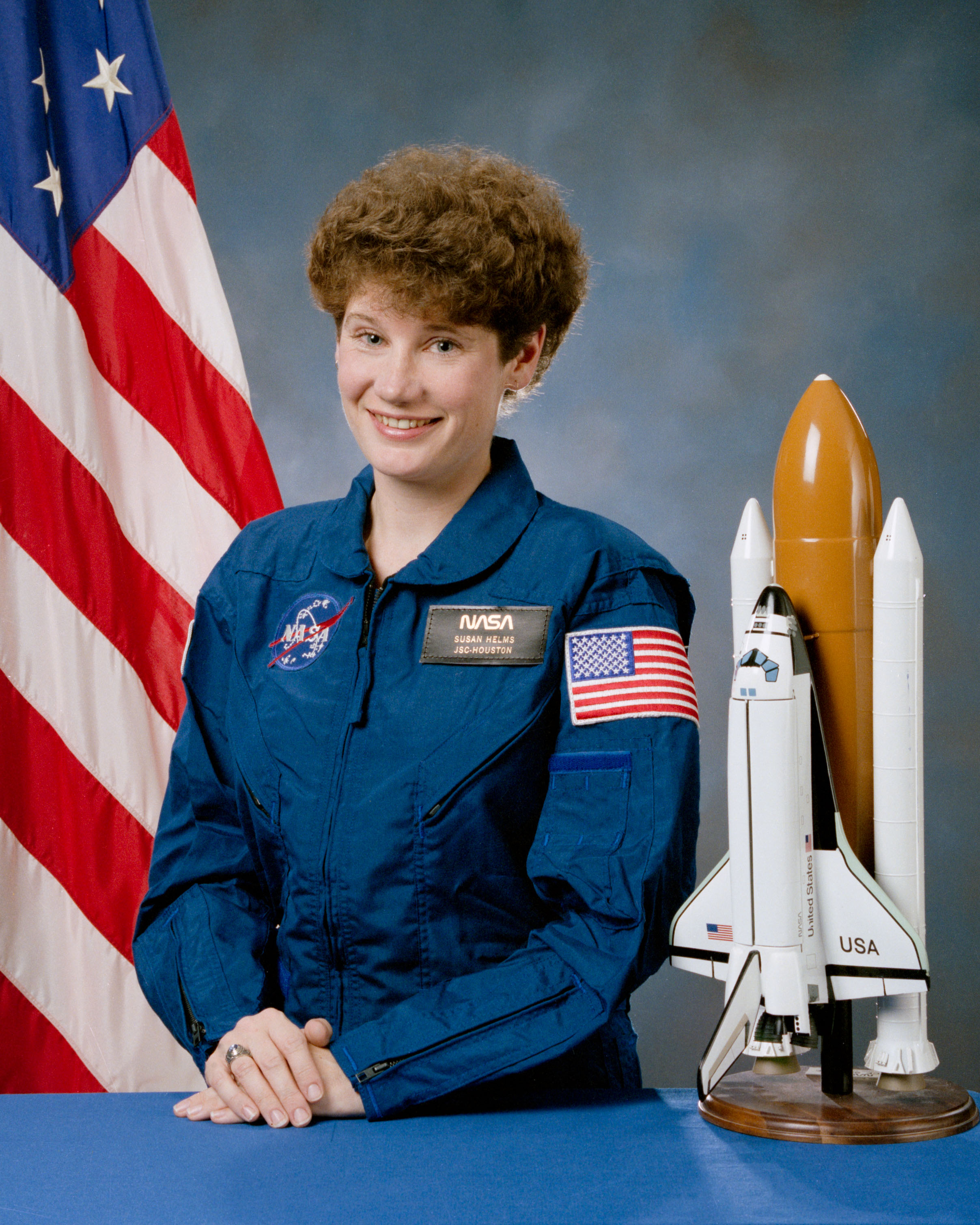 Portrait of a woman wearing a blue jumpsuit with a partially visible US flag in the background.