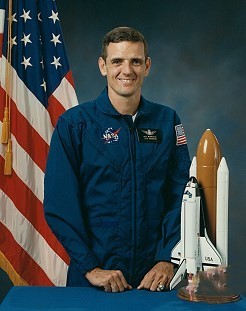 Portrait of a man wearing a blue jumpsuit with a partially visible US flag in the background.