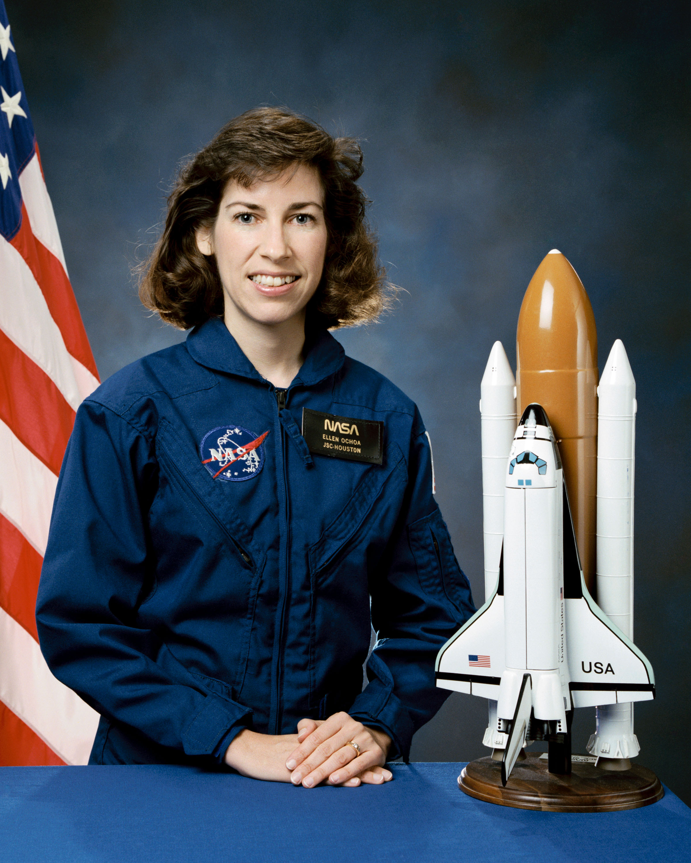 Portrait of a woman wearing a blue jumpsuit with a partially visible US flag in the background.