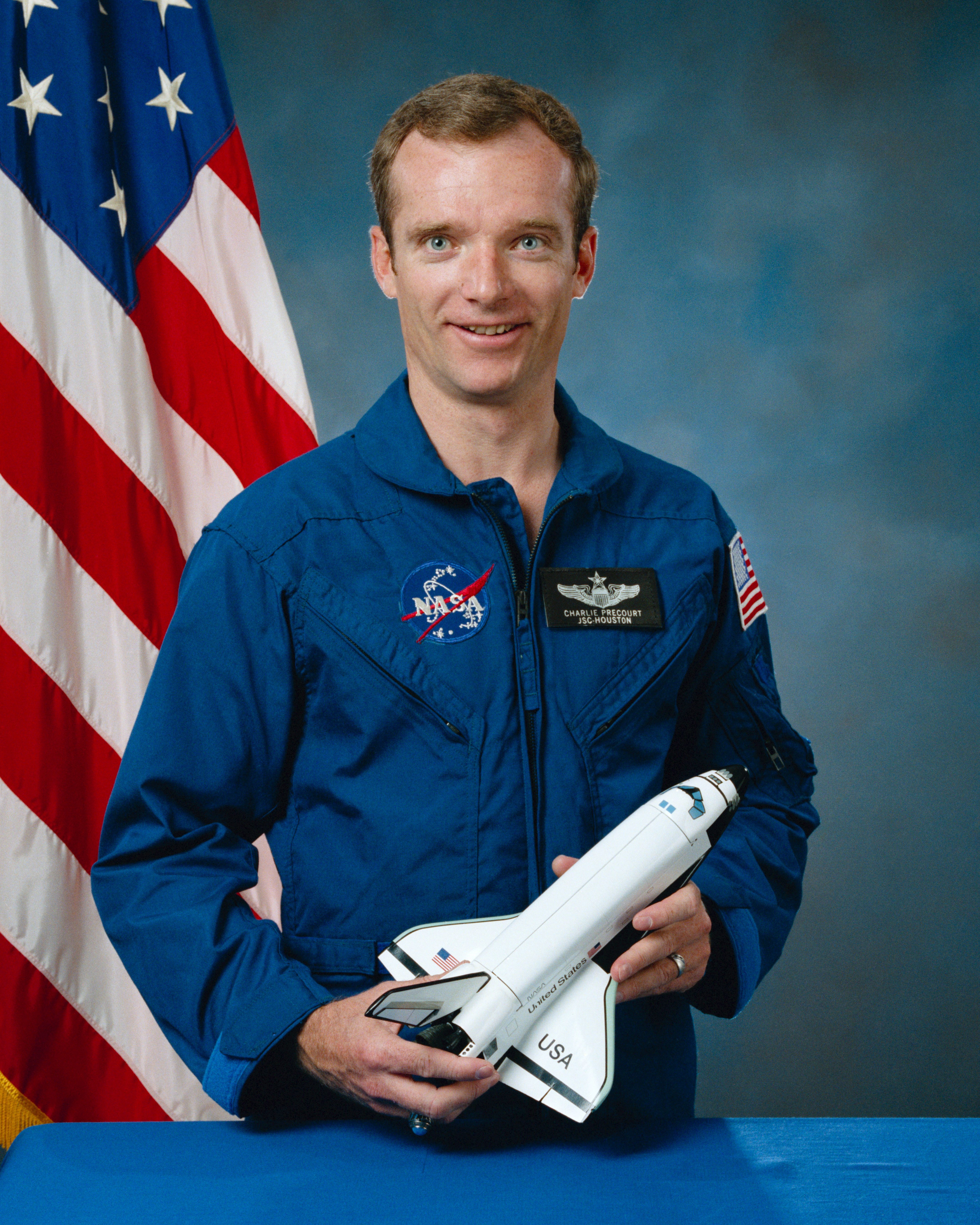 Portrait of a man wearing a blue jumpsuit with a partially visible US flag in the background.