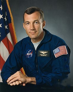 Portrait of a man wearing a blue jumpsuit with a partially visible US flag in the background.