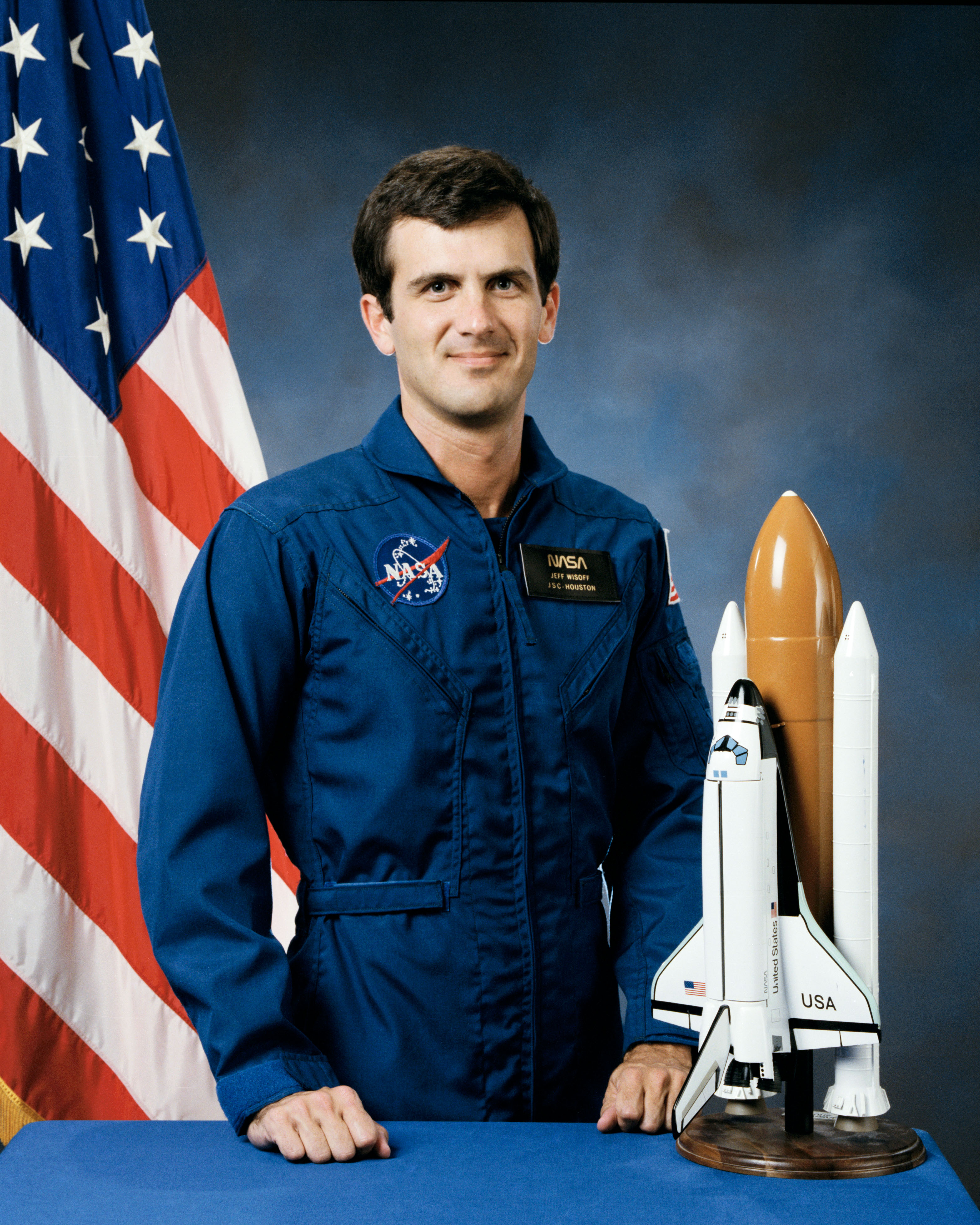 Portrait of a man wearing a blue jumpsuit with a partially visible US flag in the background.