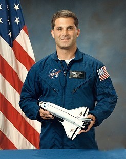 Portrait of a man wearing a blue jumpsuit with a partially visible US flag in the background.