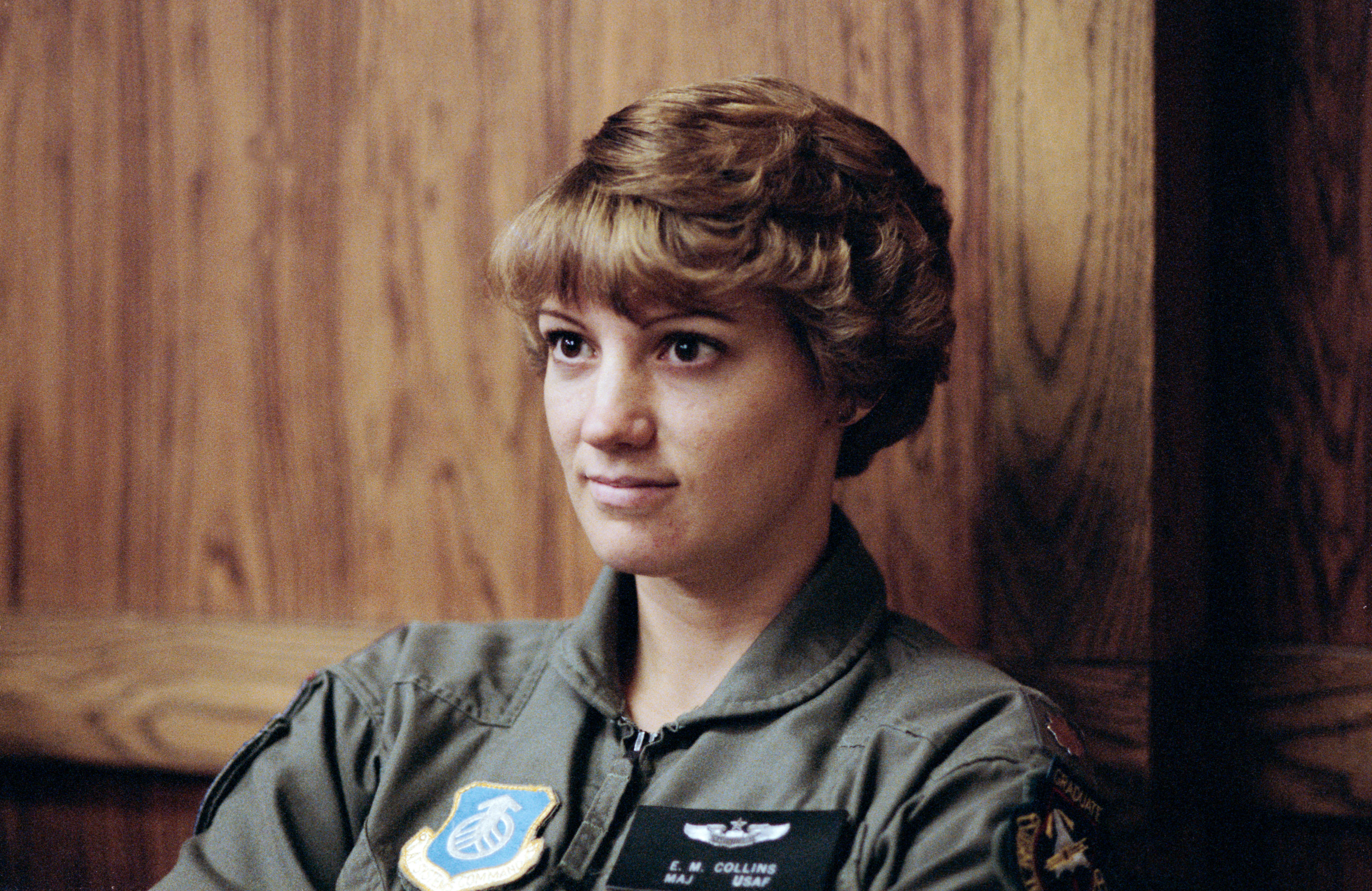 A woman wearing olive drab sits against a wood paneled wall