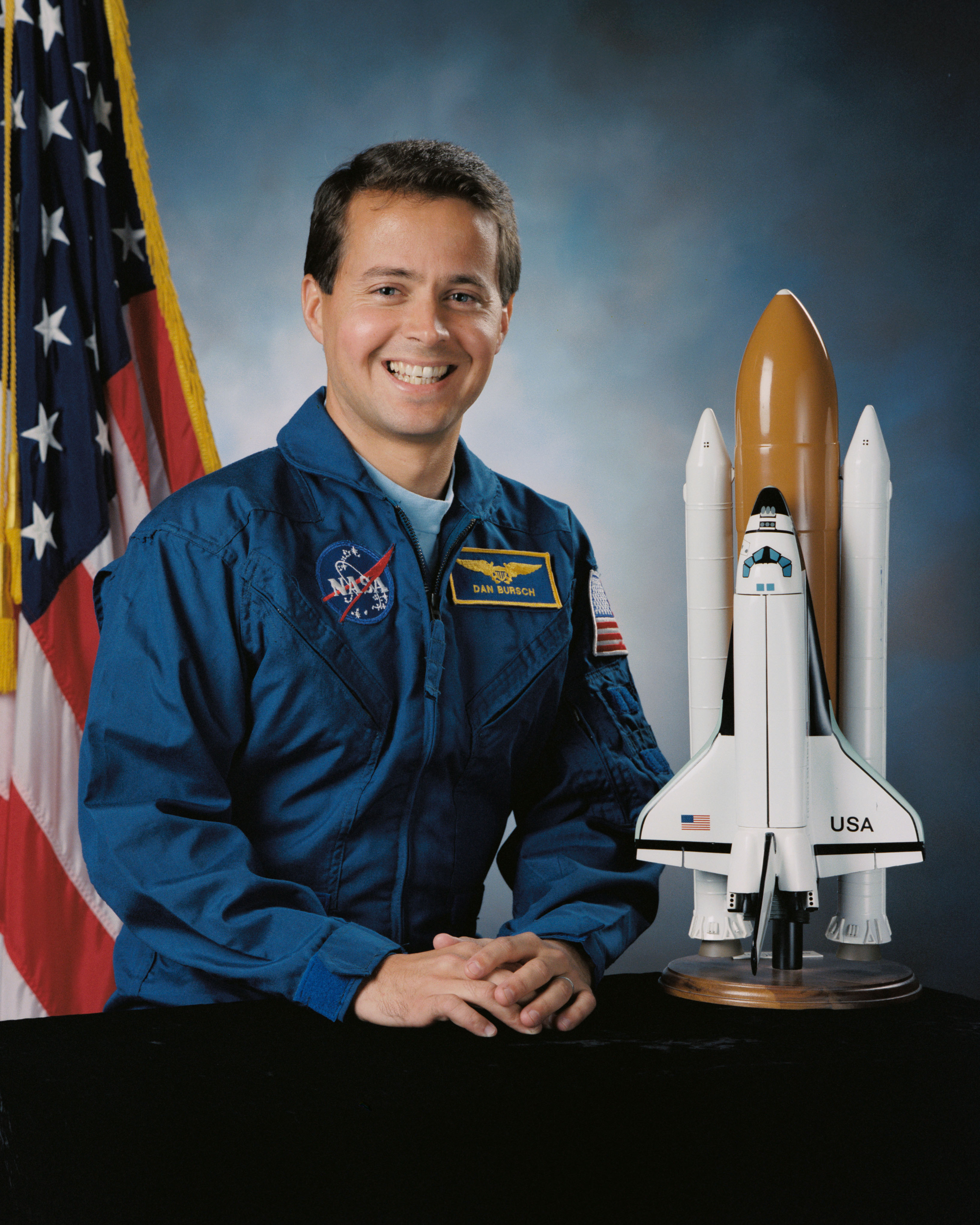 Portrait of a man wearing a blue jump suit with a partially visible US flag in the background and a model of a space shuttle