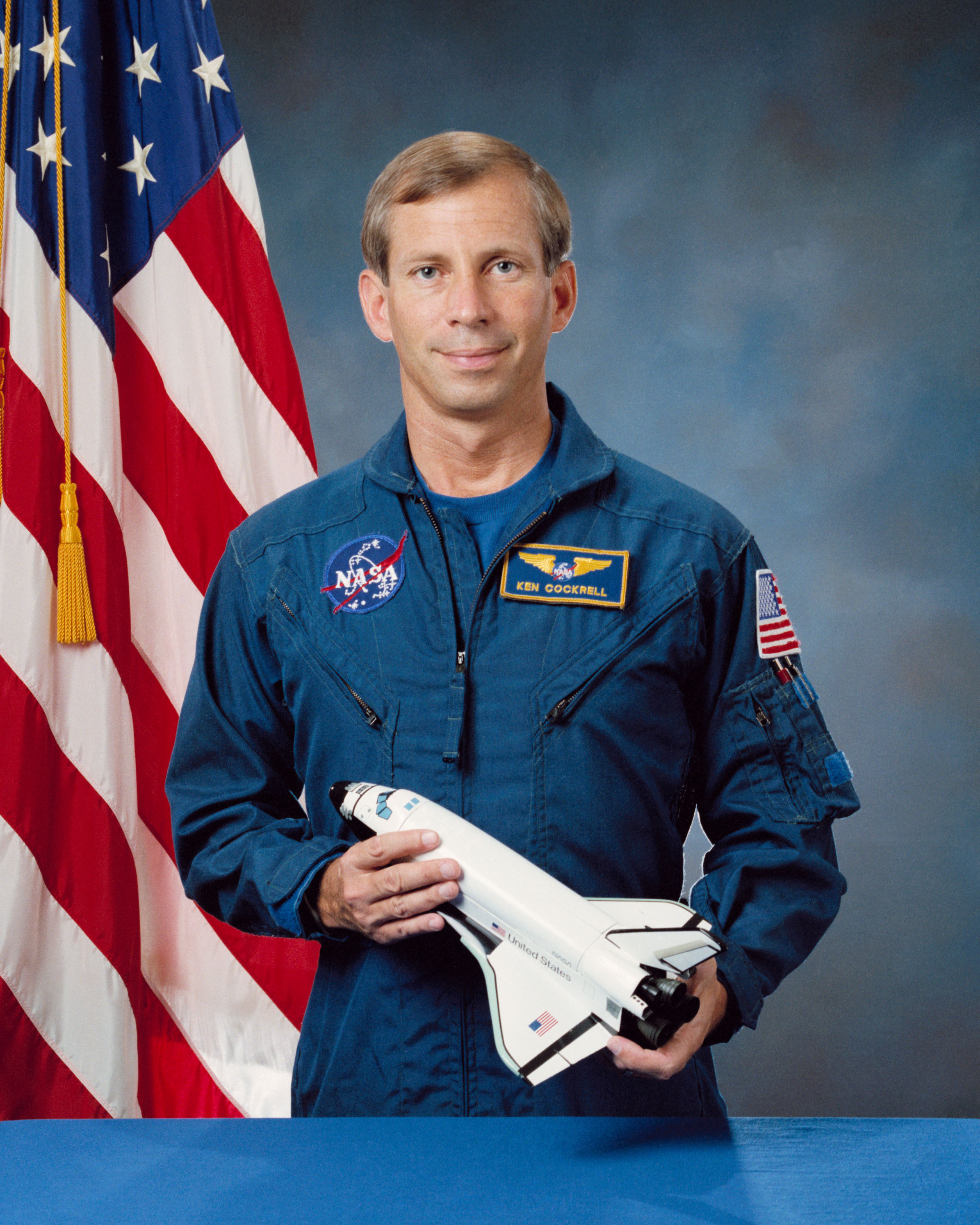 Portrait of a man wearing a blue jumpsuit with a partially visible US flag in the background