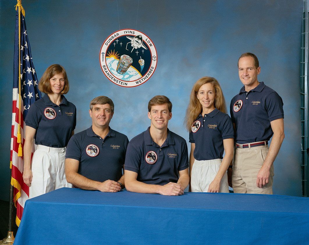 Group photo of two women and three men wearing matching blue polo shirts, with a draped American flag on the left and a round crew patch above their heads in the background.