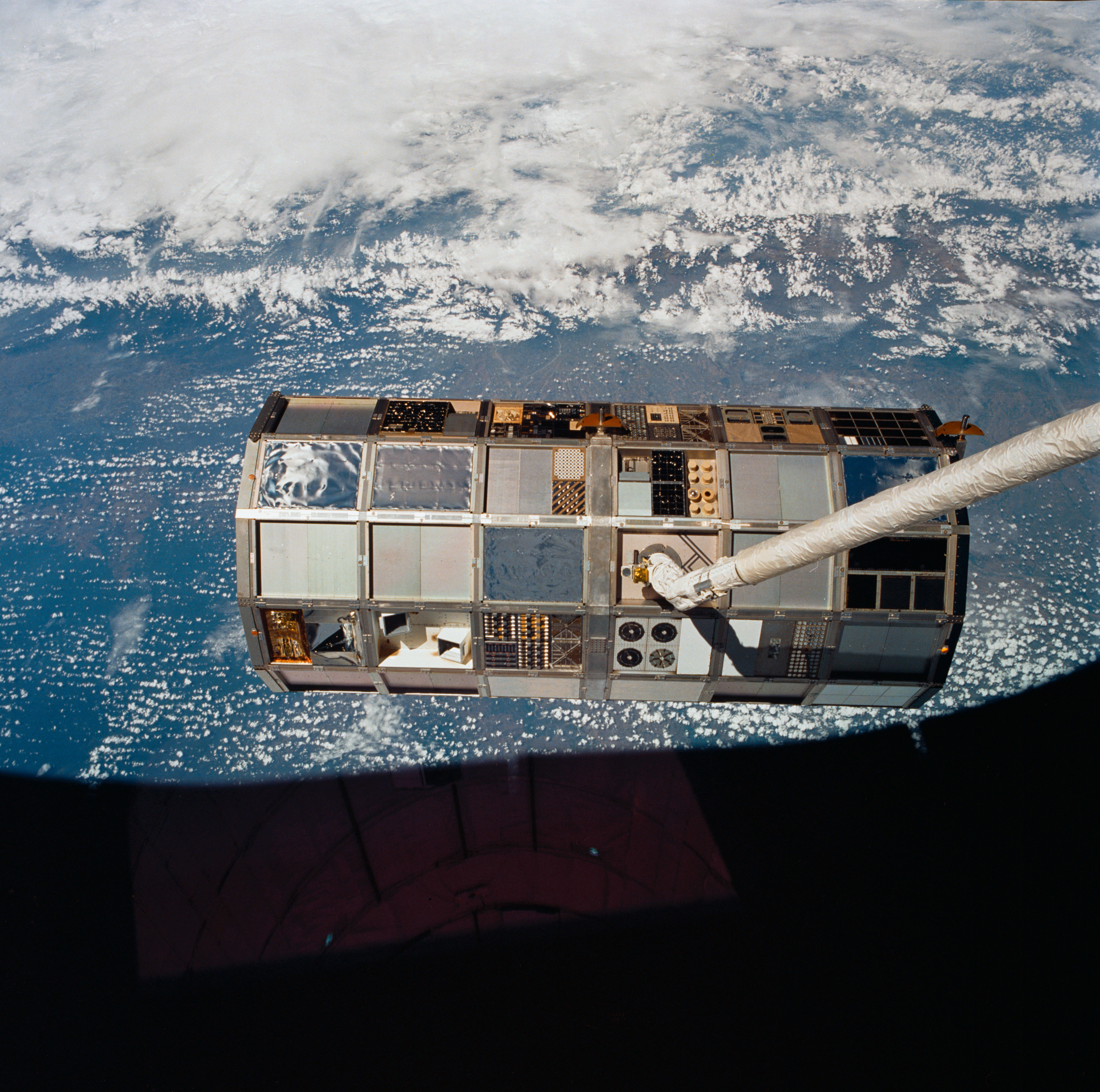 The large roughly cylindrical satellite is seen edge on, with a white robotic arm grappling it, against the blue and white limb of the Earth