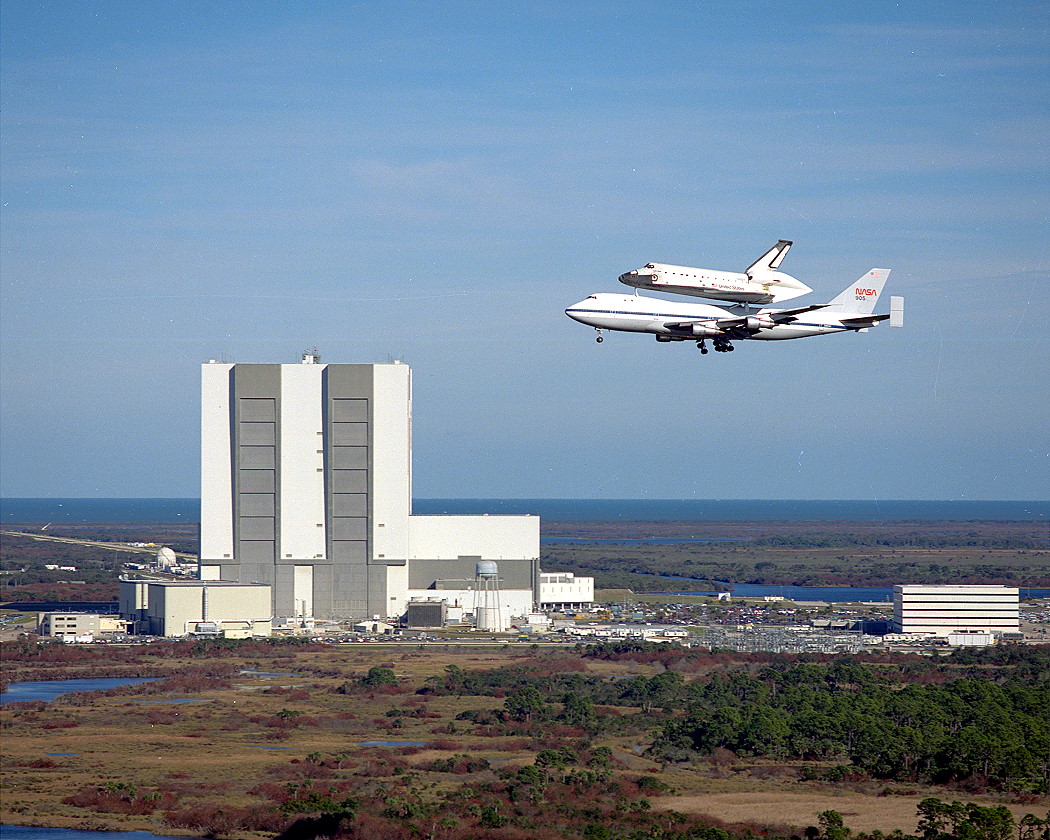 A large airplane (Boeing-747) with a space shuttle on top flies over a large building on its way to a touchdown.