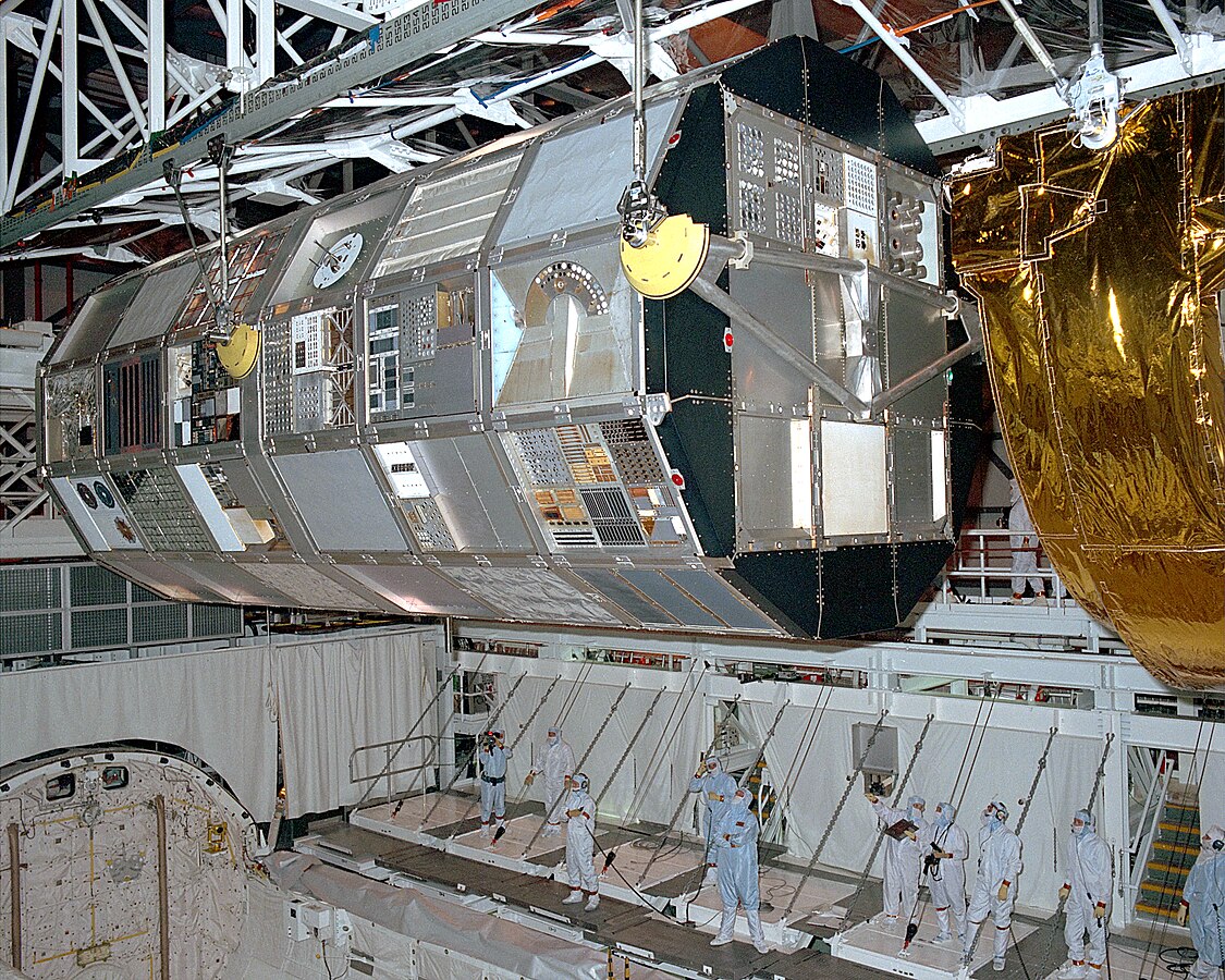 In a ground-based hanger, a large nearly cylindrical object (a satellite) is being lifted out of the space shuttle's cargo bay