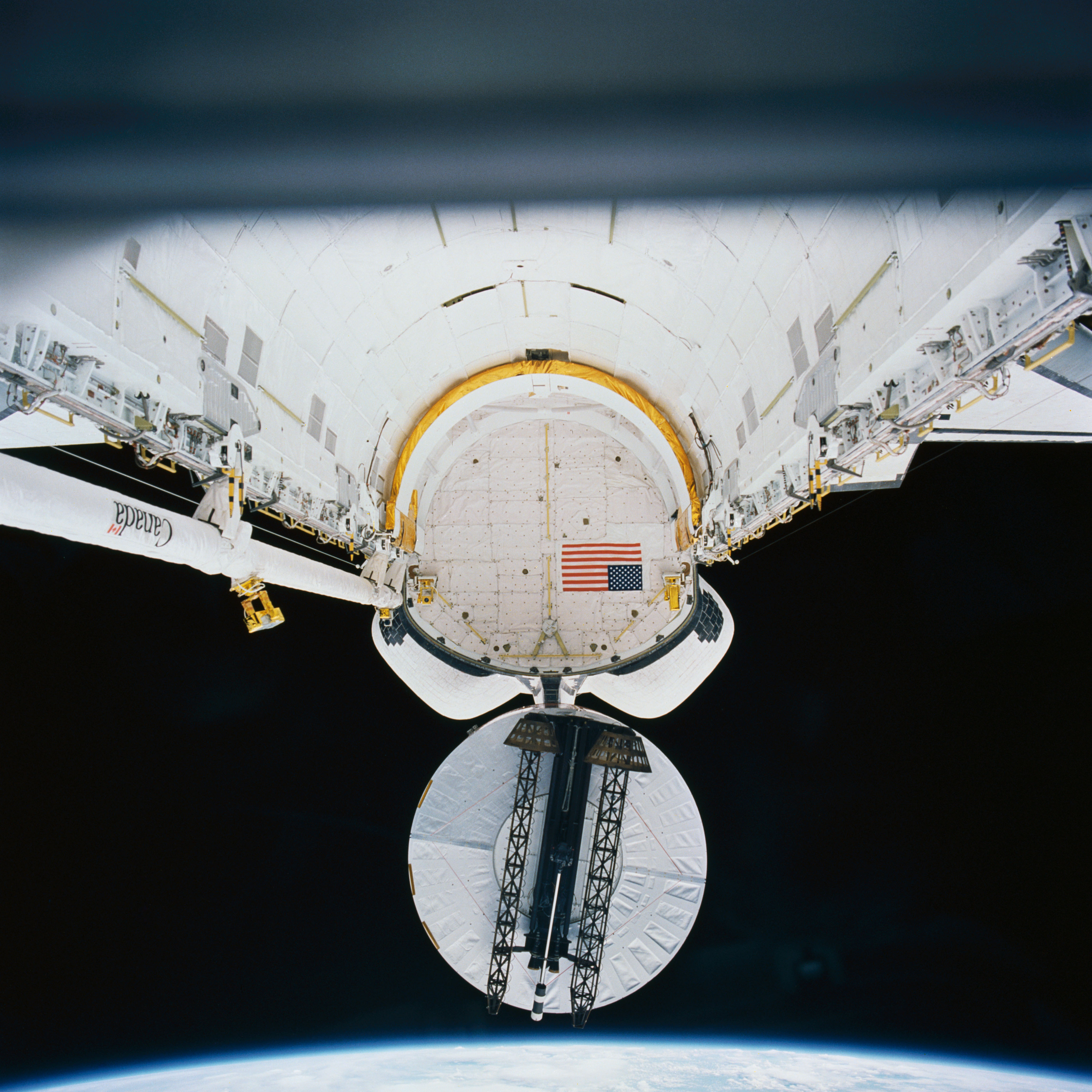 Upside down view of a space shuttle payload bay with a round white satellite emerging from it toward the bottom of the image, against a black background.